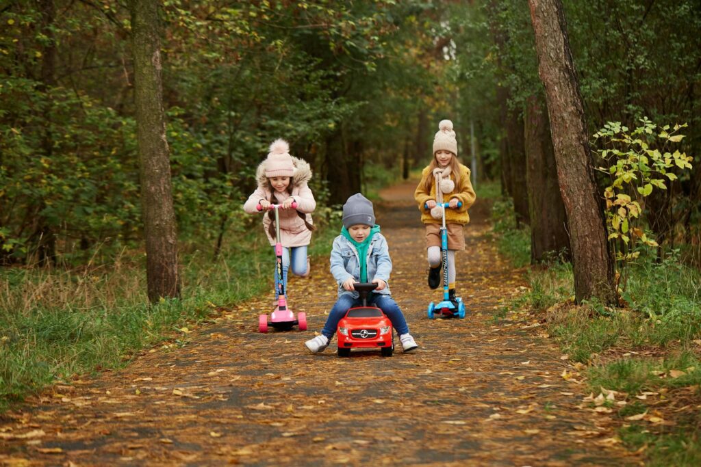 Three children enjoy a playful ride with scooters and a toy car on a forest pathway in autumn.